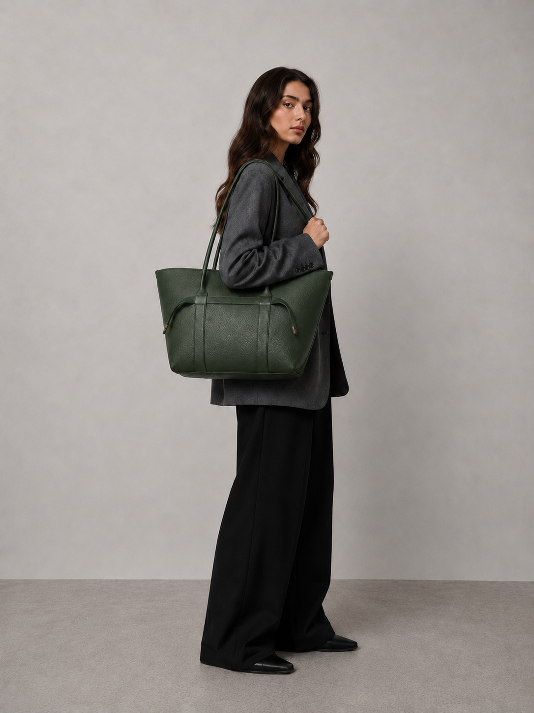 A woman in a dark gray blazer and black wide-leg trousers carries a large dark green leather tote bag with gold-tone hardware, standing against a neutral gray studio backdrop.